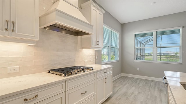 kitchen featuring baseboards, custom range hood, stainless steel gas stovetop, white cabinetry, and backsplash