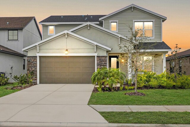 traditional-style house with a shingled roof, an attached garage, stone siding, a yard, and driveway