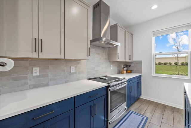 kitchen featuring blue cabinetry, stainless steel range with electric stovetop, light countertops, baseboards, and wall chimney exhaust hood