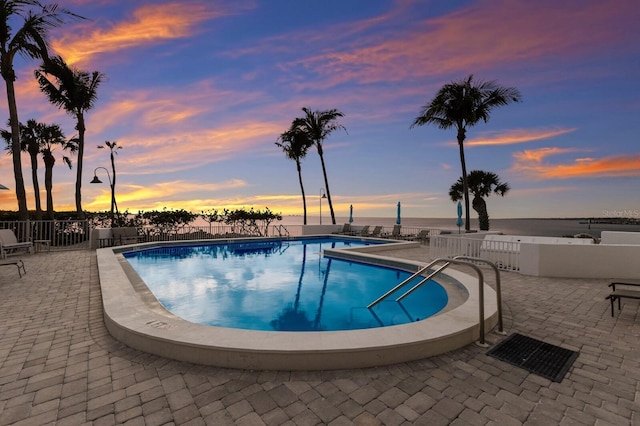 pool at dusk with a water view, a patio area, a community pool, and fence