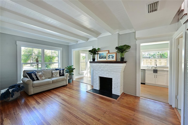 living area with light wood-style flooring, beamed ceiling, a brick fireplace, and visible vents