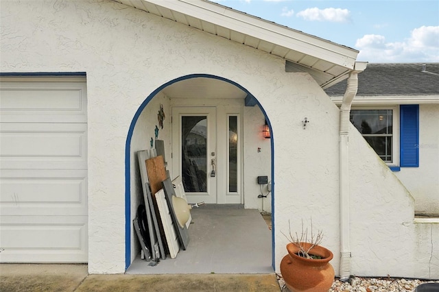 view of exterior entry featuring an attached garage, roof with shingles, and stucco siding