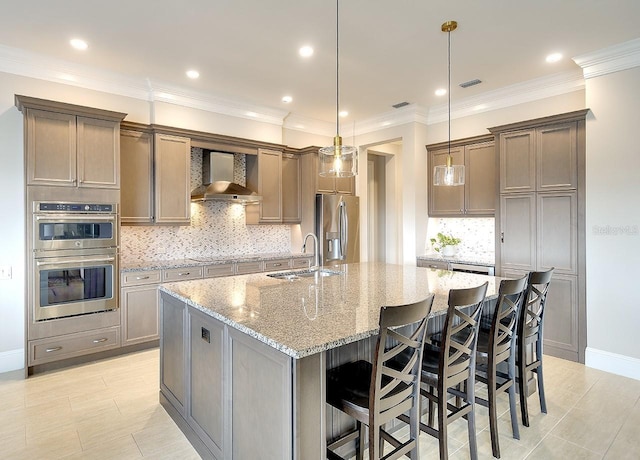 kitchen with an island with sink, light stone countertops, stainless steel appliances, wall chimney range hood, and a sink