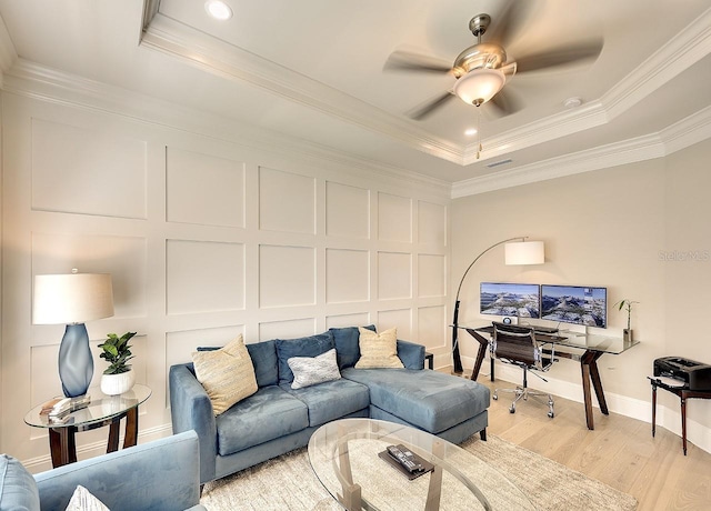 living room with light wood finished floors, a ceiling fan, ornamental molding, a tray ceiling, and a decorative wall