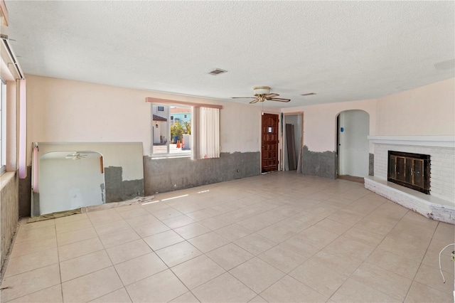 unfurnished living room featuring light tile patterned floors, arched walkways, ceiling fan, a textured ceiling, and a brick fireplace