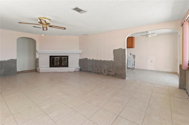 unfurnished living room with arched walkways, a textured ceiling, visible vents, a ceiling fan, and a brick fireplace