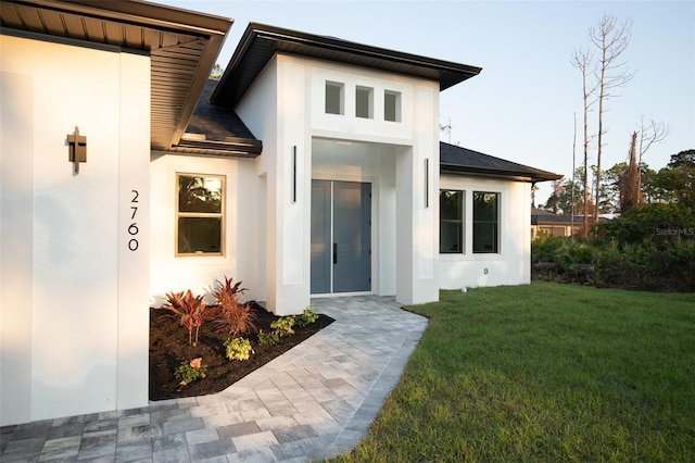 view of exterior entry featuring stucco siding, a shingled roof, and a yard