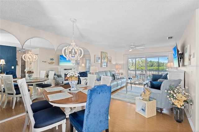 dining area with visible vents, wood finished floors, a lit fireplace, a textured ceiling, and ceiling fan with notable chandelier