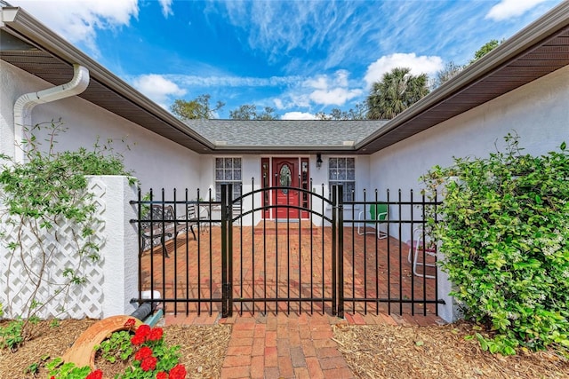 view of exterior entry with roof with shingles, a gate, fence, and stucco siding