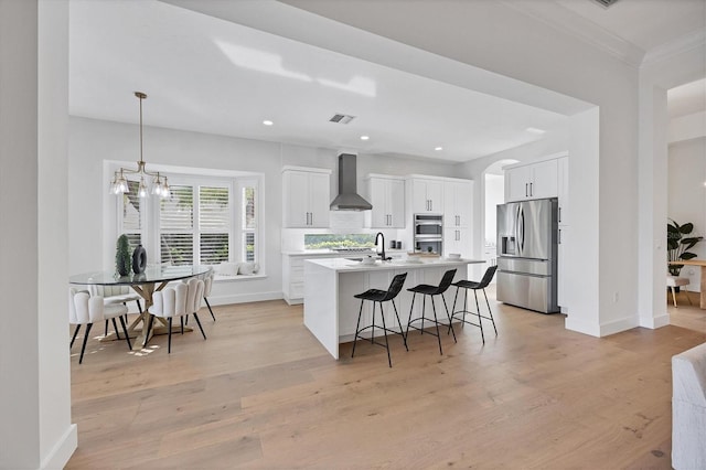 kitchen with visible vents, light wood-style floors, wall chimney range hood, stainless steel refrigerator with ice dispenser, and a kitchen bar