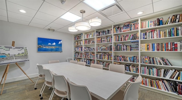 carpeted office space featuring baseboards, a drop ceiling, and wall of books