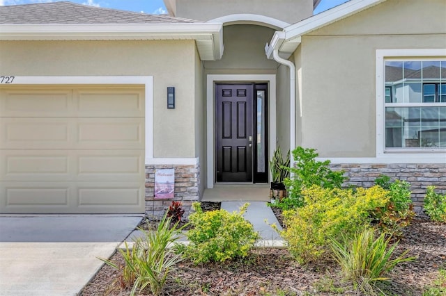 view of exterior entry with an attached garage, stone siding, a shingled roof, and stucco siding