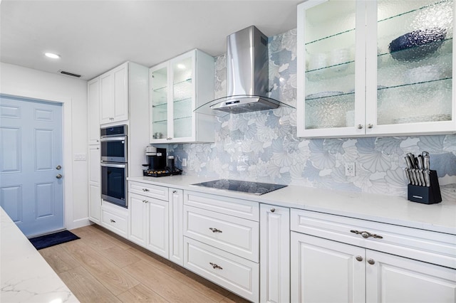 kitchen featuring light countertops, light wood-style flooring, stainless steel double oven, wall chimney range hood, and black electric cooktop