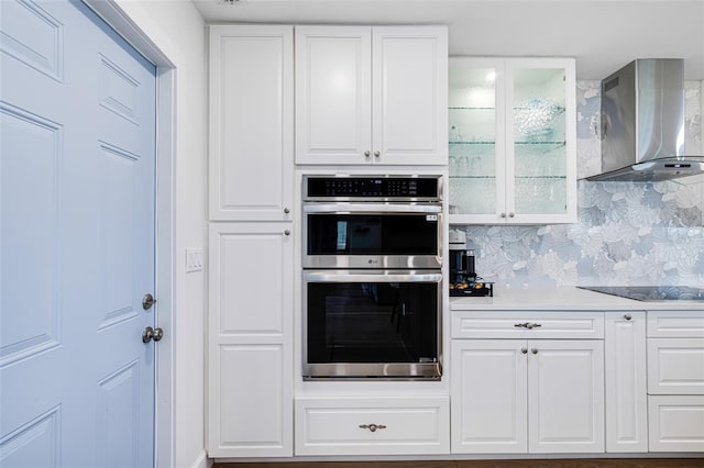 kitchen featuring black electric cooktop, stainless steel double oven, wall chimney range hood, white cabinetry, and backsplash