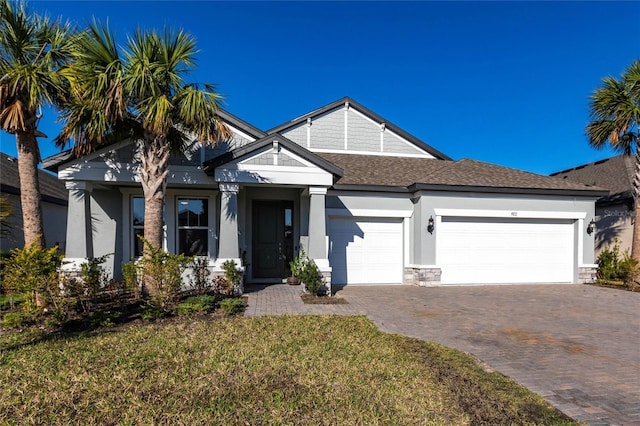 craftsman inspired home with decorative driveway, stucco siding, a shingled roof, an attached garage, and a front lawn