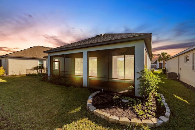 back of property at dusk featuring a sunroom, stucco siding, a lawn, and central air condition unit