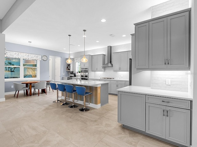 kitchen featuring a breakfast bar area, a kitchen island with sink, gray cabinets, light countertops, and wall chimney exhaust hood