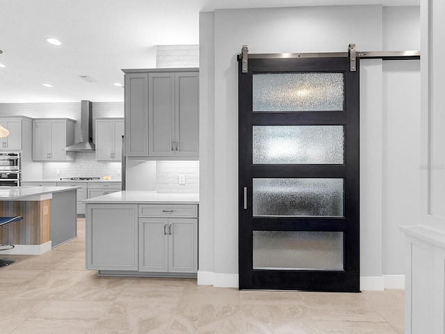 kitchen with gray cabinetry, light countertops, a barn door, wall chimney exhaust hood, and backsplash