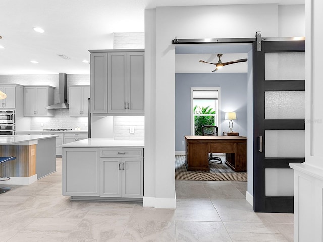 kitchen featuring gray cabinetry, wall chimney range hood, backsplash, a barn door, and light countertops