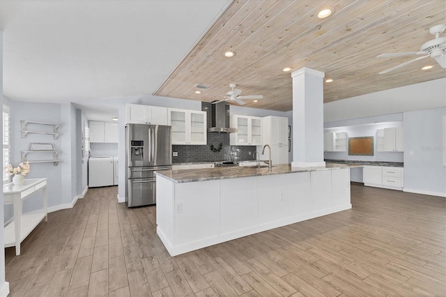 kitchen featuring backsplash, a ceiling fan, dark stone countertops, stainless steel fridge, and wall chimney exhaust hood