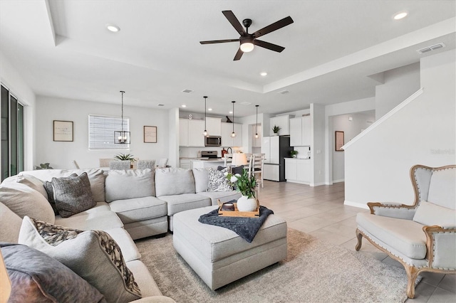 living room with ceiling fan with notable chandelier, a tray ceiling, light tile patterned flooring, and recessed lighting