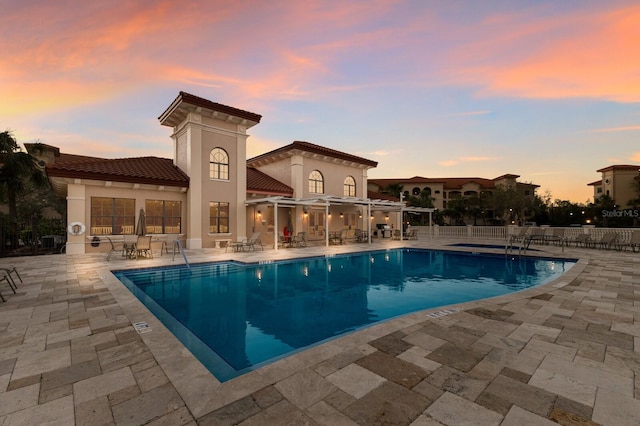 pool at dusk with a patio area, fence, and a community pool