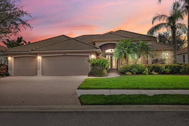 mediterranean / spanish house with a garage, a lawn, a tiled roof, decorative driveway, and stucco siding