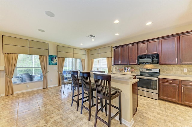 kitchen with black microwave, a kitchen island with sink, light countertops, stainless steel electric range, and decorative backsplash