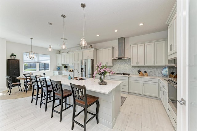 kitchen featuring a kitchen island with sink, stainless steel appliances, a sink, backsplash, and wall chimney exhaust hood