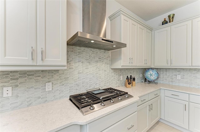 kitchen featuring wall chimney exhaust hood, light stone counters, stainless steel gas stovetop, white cabinetry, and backsplash