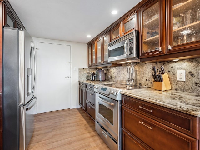 kitchen with light stone counters, decorative backsplash, appliances with stainless steel finishes, glass insert cabinets, and light wood-type flooring