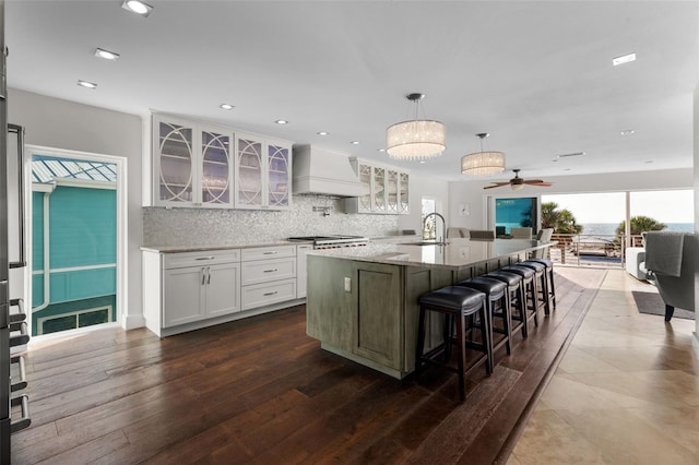 kitchen featuring white cabinets, backsplash, a kitchen island with sink, premium range hood, and a sink