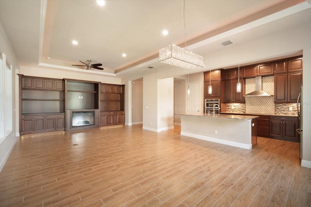 kitchen featuring light wood finished floors, a center island with sink, a raised ceiling, and wall chimney range hood