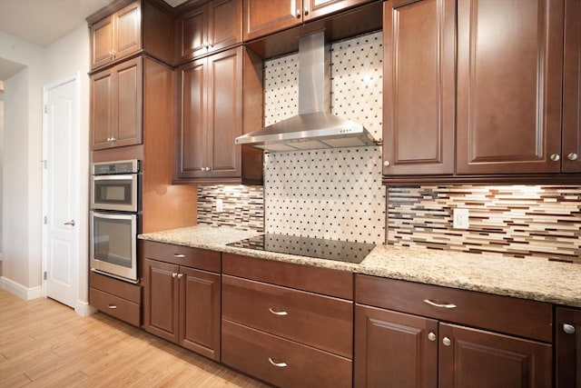 kitchen with light wood finished floors, light stone countertops, decorative backsplash, stainless steel double oven, and wall chimney exhaust hood