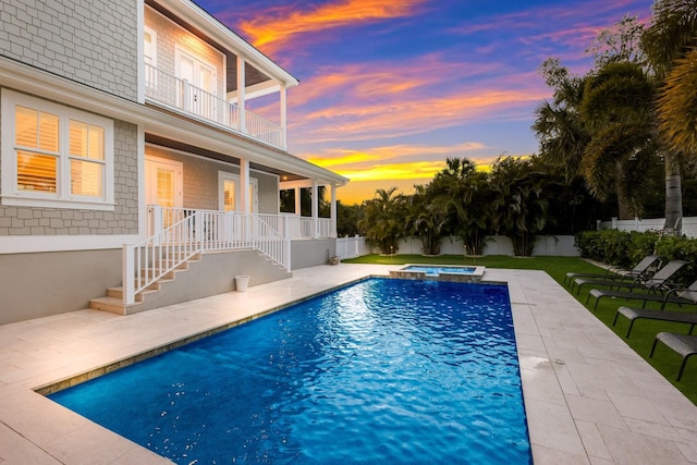 pool at dusk with a patio area, a pool with connected hot tub, fence, and a lawn