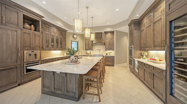 kitchen featuring light stone countertops, a center island with sink, stainless steel double oven, decorative backsplash, and custom range hood