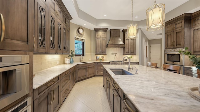 kitchen with tasteful backsplash, premium range hood, decorative light fixtures, light stone counters, and a sink