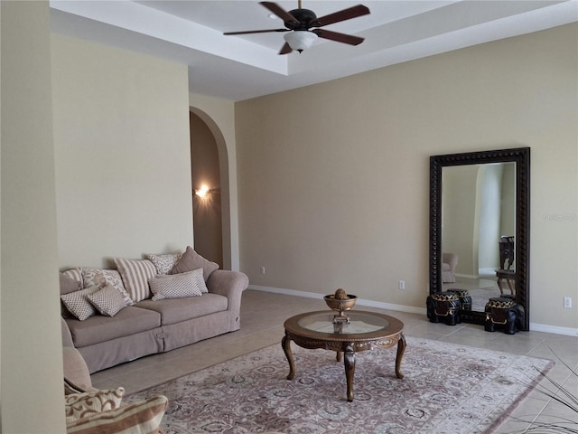 tiled living room featuring arched walkways, a raised ceiling, ceiling fan, and baseboards