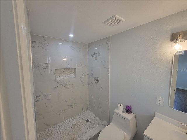 bathroom with visible vents, tiled shower, a textured wall, toilet, and vanity