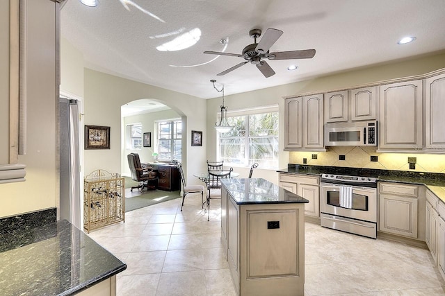 kitchen featuring arched walkways, light tile patterned floors, appliances with stainless steel finishes, a kitchen island, and dark stone countertops