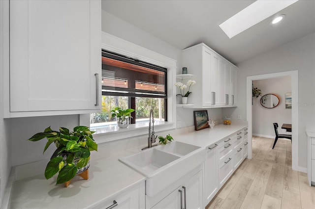 kitchen featuring light wood-type flooring, light countertops, vaulted ceiling with skylight, white cabinets, and a sink