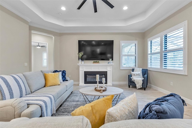 living room featuring a tray ceiling, a glass covered fireplace, ceiling fan, and baseboards