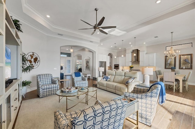 living room featuring a tray ceiling, arched walkways, light wood-style flooring, ornamental molding, and ceiling fan with notable chandelier