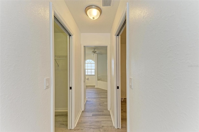 hallway featuring light wood-type flooring, baseboards, visible vents, and a textured wall