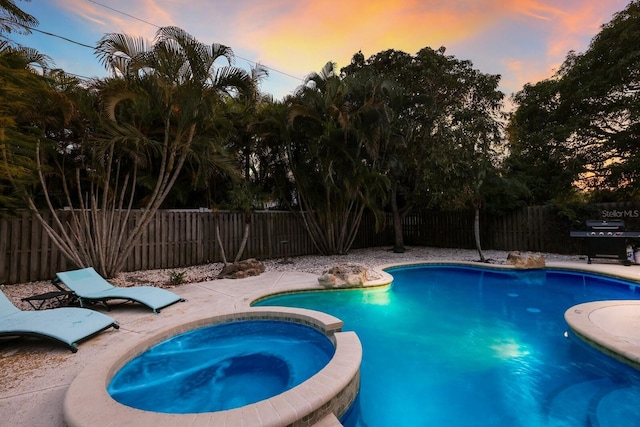 pool at dusk with a pool with connected hot tub and a fenced backyard