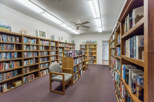 sitting room featuring a textured ceiling, wall of books, carpet flooring, and a ceiling fan