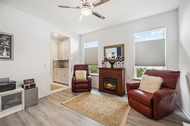 sitting room featuring light wood-style floors, a warm lit fireplace, baseboards, and a ceiling fan