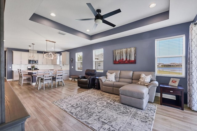 living room with light wood-style flooring, visible vents, a tray ceiling, and a ceiling fan