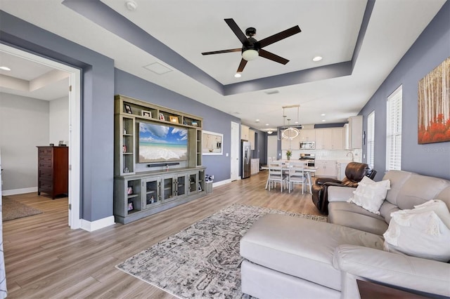 living room featuring a tray ceiling, light wood-type flooring, a ceiling fan, and baseboards