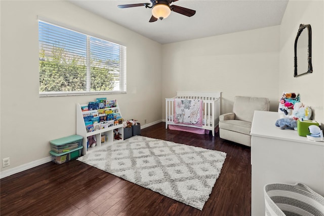 bedroom with a nursery area, dark wood finished floors, a ceiling fan, and baseboards